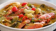 A close-up of a dish featuring seafood, including crab legs, shrimp, clams, and lobster. The dish is garnished with chopped herbs and slices of red and yellow bell peppers. The food appears to be cooked in a flavorful, thick sauce, likely containing spices and herbs.