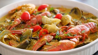 A close-up of a dish featuring seafood, including crab legs, shrimp, clams, and lobster. The dish is garnished with chopped herbs and slices of red and yellow bell peppers. The food appears to be cooked in a flavorful, thick sauce, likely containing spices and herbs.