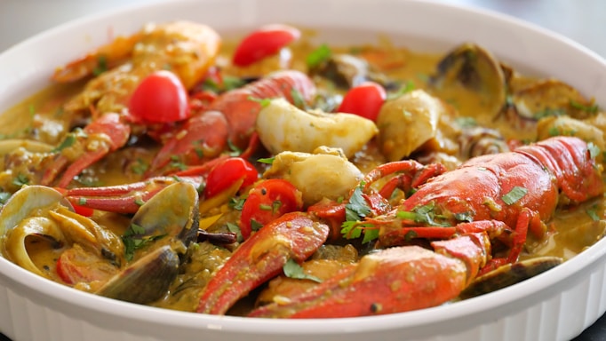 A close-up of a dish featuring seafood, including crab legs, shrimp, clams, and lobster. The dish is garnished with chopped herbs and slices of red and yellow bell peppers. The food appears to be cooked in a flavorful, thick sauce, likely containing spices and herbs.
