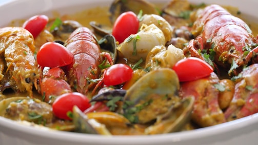A colorful seafood dish featuring large pieces of lobster or crab, clams, and scallops mixed with cherry tomatoes and garnished with fresh herbs. The dish is presented in a rich, creamy sauce.