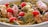 A colorful seafood dish featuring large pieces of lobster or crab, clams, and scallops mixed with cherry tomatoes and garnished with fresh herbs. The dish is presented in a rich, creamy sauce.