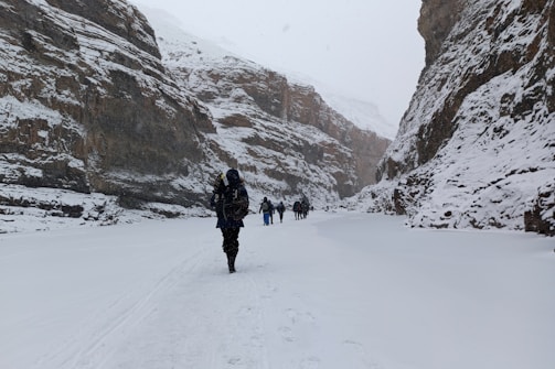 A group of people is trekking through a deep snow-covered valley flanked by towering rock walls. The ground and cliffs are blanketed in snow, indicating a cold and wintry environment. People are dressed in heavy winter clothing, suggesting harsh weather conditions.