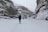 A group of people is trekking through a deep snow-covered valley flanked by towering rock walls. The ground and cliffs are blanketed in snow, indicating a cold and wintry environment. People are dressed in heavy winter clothing, suggesting harsh weather conditions.