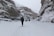A group of people is trekking through a deep snow-covered valley flanked by towering rock walls. The ground and cliffs are blanketed in snow, indicating a cold and wintry environment. People are dressed in heavy winter clothing, suggesting harsh weather conditions.