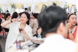 A woman is making a peace sign with her fingers while holding a glass of a dark beverage. She is smiling and appears to be enjoying herself at a social gathering or party. The background shows other people socializing at tables under a white and yellow canopy. On the table in front of her, there is a Pepsi can, a dish of food, and a water bottle.