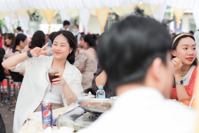 A woman is making a peace sign with her fingers while holding a glass of a dark beverage. She is smiling and appears to be enjoying herself at a social gathering or party. The background shows other people socializing at tables under a white and yellow canopy. On the table in front of her, there is a Pepsi can, a dish of food, and a water bottle.