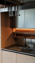 A modern kitchen sink area with wooden cabinetry and a sleek metal faucet. The minimalist design includes overhead shelving with warm ambient lighting underneath.