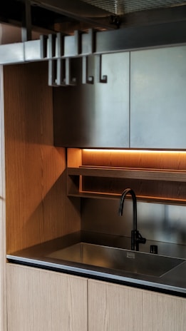 A modern kitchen sink area with wooden cabinetry and a sleek metal faucet. The minimalist design includes overhead shelving with warm ambient lighting underneath.