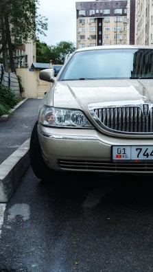 A beige car is parked on a wet street near a curb in an urban setting. The car is wet with raindrops, suggesting recent rain. In the background, there are multi-story residential buildings and some greenery on the left side. The license plate on the car indicates a specific region.