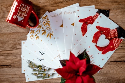 A collection of holiday greeting cards is spread out on a wooden surface. The cards feature festive designs, including red mittens holding a heart-shaped snowball and golden leaves. A red mug with winter-themed illustrations and a poinsettia plant add a seasonal touch.