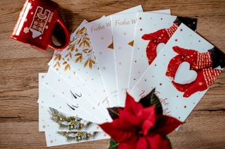 A collection of holiday greeting cards is spread out on a wooden surface. The cards feature festive designs, including red mittens holding a heart-shaped snowball and golden leaves. A red mug with winter-themed illustrations and a poinsettia plant add a seasonal touch.