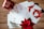 A collection of holiday greeting cards is spread out on a wooden surface. The cards feature festive designs, including red mittens holding a heart-shaped snowball and golden leaves. A red mug with winter-themed illustrations and a poinsettia plant add a seasonal touch.
