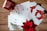 A collection of holiday greeting cards is spread out on a wooden surface. The cards feature festive designs, including red mittens holding a heart-shaped snowball and golden leaves. A red mug with winter-themed illustrations and a poinsettia plant add a seasonal touch.