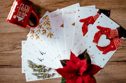 A collection of holiday greeting cards is spread out on a wooden surface. The cards feature festive designs, including red mittens holding a heart-shaped snowball and golden leaves. A red mug with winter-themed illustrations and a poinsettia plant add a seasonal touch.
