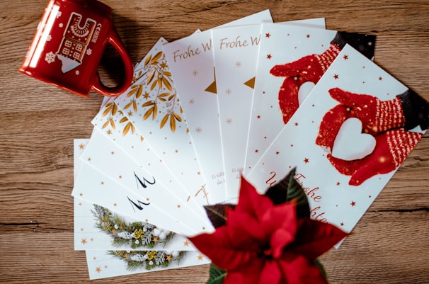 A collection of holiday greeting cards is spread out on a wooden surface. The cards feature festive designs, including red mittens holding a heart-shaped snowball and golden leaves. A red mug with winter-themed illustrations and a poinsettia plant add a seasonal touch.