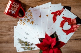 A collection of holiday greeting cards is spread out on a wooden surface. The cards feature festive designs, including red mittens holding a heart-shaped snowball and golden leaves. A red mug with winter-themed illustrations and a poinsettia plant add a seasonal touch.