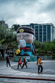 A large, colorful statue resembling a traditional Chinese figure with a white beard and mustache, holding a gold ingot, stands prominently in an urban area with modern buildings in the background. Several people walk by on the paved plaza in front of the statue, some wearing coats and masks.
