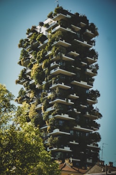 A modern skyscraper adorned with lush greenery on its balconies, creating a vertical forest-like appearance. The building's design integrates plants and trees into its architecture, making it stand out against a clear blue sky. Surrounding the structure are additional trees, contributing to the verdant atmosphere.