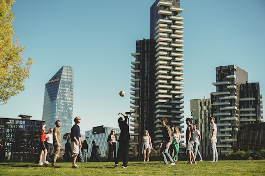 A group of people enjoys playing volleyball on a grassy area surrounded by tall modern buildings. The participants appear relaxed and engaged in the activity amidst the urban backdrop, with clear blue skies above.