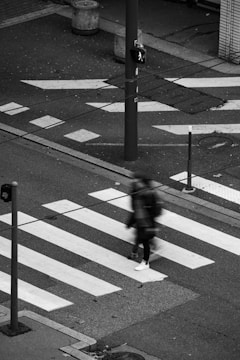 A person is crossing a zebra-striped pedestrian crosswalk on a street. It appears to be an urban setting with a visible pedestrian signal lit up, indicating it is safe to walk. The image is in black and white, adding a sense of timelessness and moodiness. There are also several street poles and a pedestrian crossing sign visible.