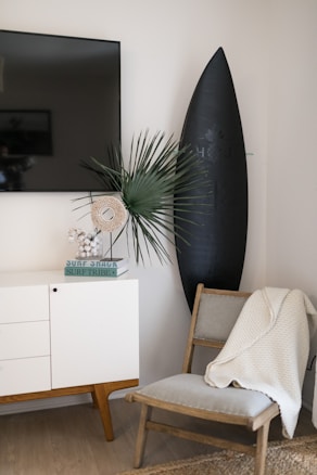A modern, minimalist living space features a wooden chair draped with a white knitted blanket beside a sleek, black surfboard with a logo. A white cabinet with mid-century style legs is topped with books, a decorative piece containing small balls, and a green plant with spiky leaves. A flat-screen TV is mounted on the wall above, providing a contemporary vibe.