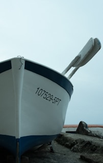 A white boat with a blue trim is positioned on land next to some large rocks. The registration number 107529-5PT is visible on the front side of the boat. A pair of oars is affixed to the boat. The background appears to be an overcast sky with a hint of water or horizon in the distance.
