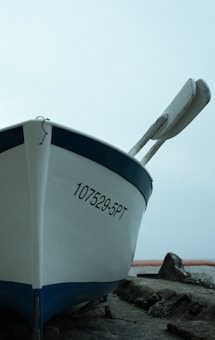 A white boat with a blue trim is positioned on land next to some large rocks. The registration number 107529-5PT is visible on the front side of the boat. A pair of oars is affixed to the boat. The background appears to be an overcast sky with a hint of water or horizon in the distance.