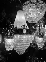 A stunning display of opulent crystal chandeliers illuminated against a dark background. The intricate designs feature numerous hanging crystals that reflect the light, creating a shimmering and luxurious atmosphere. The craftsmanship is evident in the detailed metalwork holding the crystals in place.