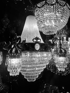 A stunning display of opulent crystal chandeliers illuminated against a dark background. The intricate designs feature numerous hanging crystals that reflect the light, creating a shimmering and luxurious atmosphere. The craftsmanship is evident in the detailed metalwork holding the crystals in place.