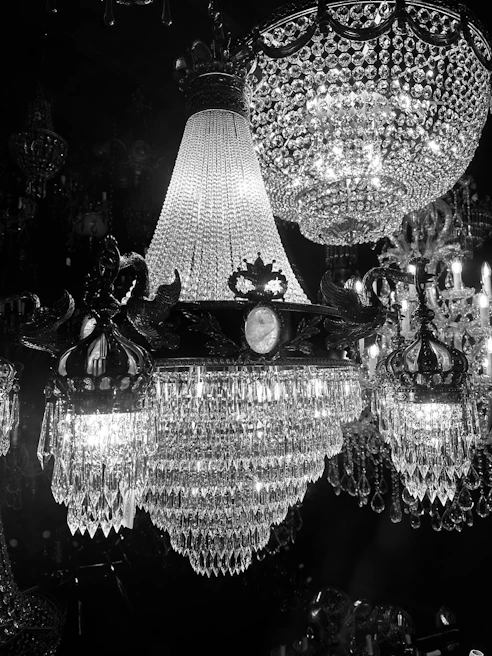 A stunning display of opulent crystal chandeliers illuminated against a dark background. The intricate designs feature numerous hanging crystals that reflect the light, creating a shimmering and luxurious atmosphere. The craftsmanship is evident in the detailed metalwork holding the crystals in place.