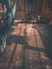 A sunlit terrace with shadows cast by railing patterns and hanging fabrics. Several potted plants are lined along the perimeter, adding greenery to the scene. The fabrics have floral and abstract designs, creating an artistic and homely atmosphere.