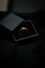 A delicate gold ring with an intricate design rests inside an open black jewelry box. The box is set against a dark, possibly velvet background, which enhances the ring's golden hue.