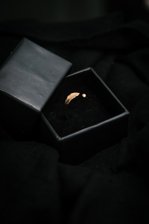 A golden ring with a small leaf design and a gemstone is placed in an open black jewelry box. The box is surrounded by a dark, soft fabric, creating a luxurious and elegant setting.