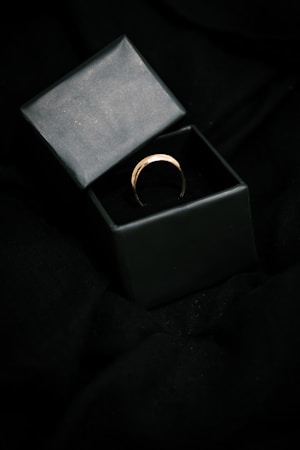A gold ring is placed inside an open black box on a dark fabric background, highlighting the elegance and simplicity of the jewelry piece.