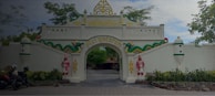 An ornate gate with white walls and decorative elements including two figures in traditional attire and a green serpent motif. The arch has gold accents and displays the text 'Puri Mataram.' Motorbikes are parked on the left side, and trees provide a natural backdrop.