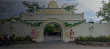 An ornate gate with white walls and decorative elements including two figures in traditional attire and a green serpent motif. The arch has gold accents and displays the text 'Puri Mataram.' Motorbikes are parked on the left side, and trees provide a natural backdrop.