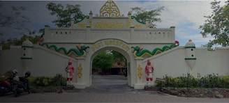 An ornate gate with white walls and decorative elements including two figures in traditional attire and a green serpent motif. The arch has gold accents and displays the text 'Puri Mataram.' Motorbikes are parked on the left side, and trees provide a natural backdrop.