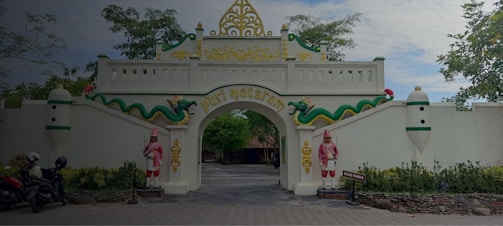 An ornate gate with white walls and decorative elements including two figures in traditional attire and a green serpent motif. The arch has gold accents and displays the text 'Puri Mataram.' Motorbikes are parked on the left side, and trees provide a natural backdrop.