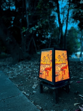 A decorative lantern featuring detailed Asian-inspired artwork with red and yellow tones stands on a small black wooden table in an outdoor setting. Surrounding the lantern are fallen leaves and dirt, with dark trees and dim lighting in the background, creating a serene ambiance.