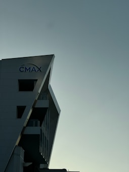 A modern building with a sharp, angular design is partially visible, with the logo 'CMAX' on the side. The background consists of a smooth, cloudy sky with dim lighting, suggesting evening or early morning.