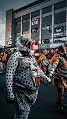 A person is dressed in an elaborate leopard costume, featuring striking artistic body paint and a detailed mask. The paint is ornately crafted with vivid colors, making the figure resemble a leopard with an open mouth and intense eyes. In the background, other people are dressed in similarly intricate animal-themed costumes, suggesting a parade or festival. A large building with various signs and banners can be seen behind the participants.