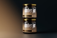 Two amber glass jars stacked vertically against a gradient background. The jars have metallic lids and labels displaying the text 'amber jar' and 'mockup templates' in bold typography. The lighting creates a soft, warm glow on the jars, highlighting their glossy surface.