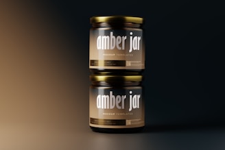 Two amber glass jars stacked vertically against a gradient background. The jars have metallic lids and labels displaying the text 'amber jar' and 'mockup templates' in bold typography. The lighting creates a soft, warm glow on the jars, highlighting their glossy surface.