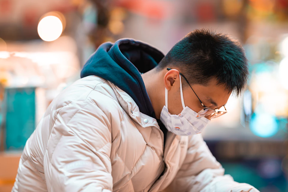 In the image, a person is seen wearing a white jacket and a face mask while sitting at a table with their head down. The background features a blurred store window filled with various items for sale, suggesting that they are likely shopping or browsing through the offerings in the store.