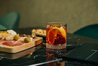 A glass filled with a red cocktail and a slice of orange sits on a black marble surface. To the left, a wooden board displays various appetizers including bread slices, green olives, cheese, and cured meats.