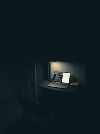 A dimly lit room features a small desk with a laptop illuminated by a desk lamp. The laptop screen displays coding text on one half and a graph or chart on the other. The room appears minimalistic with a focus on the computer setup. The ambiance is quiet and subdued, suggesting a late-night work or study session.