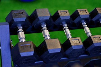 A set of hexagonal dumbbells is neatly arranged on a rack, with a visible weight measurement of 5KG. The surrounding area has a vibrant green background, suggesting the presence of artificial grass or a similar surface.