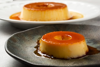 Two servings of caramel flan sit on separate plates, with their smooth, golden custard topped by a shiny layer of caramel sauce. The foreground flan is on a dark, textured plate, while the background flan is on a white plate.