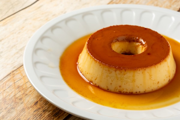 A creamy, caramel-flavored flan sits on a round white plate. The dessert has a glossy topping and is surrounded by a light layer of caramel sauce. The background features a wooden surface, adding a rustic touch.