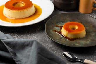 Two servings of caramel flan are presented, one on a white plate and the other on a dark, textured plate. The flans have a caramel glaze on top and are placed on a textured, dark-colored surface. Nearby are a black cloth, a small black spoon, and a ceramic cup.